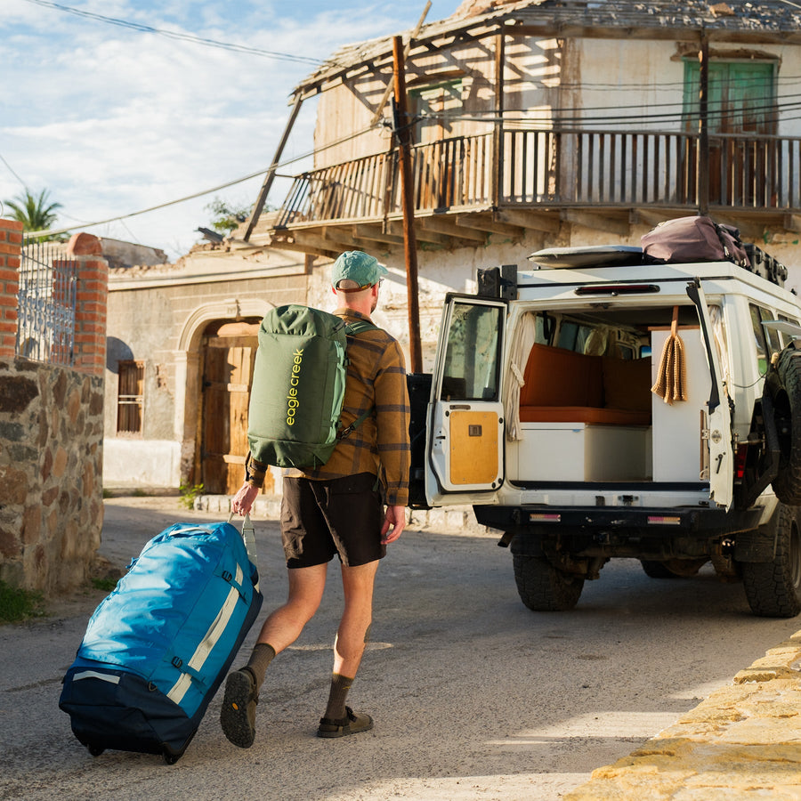 A man wearing a green duffle bag backpack wheels a large blue duffle bag to a camper van