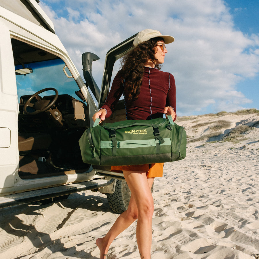A woman carrying a green duffle bag across a sandy beach
