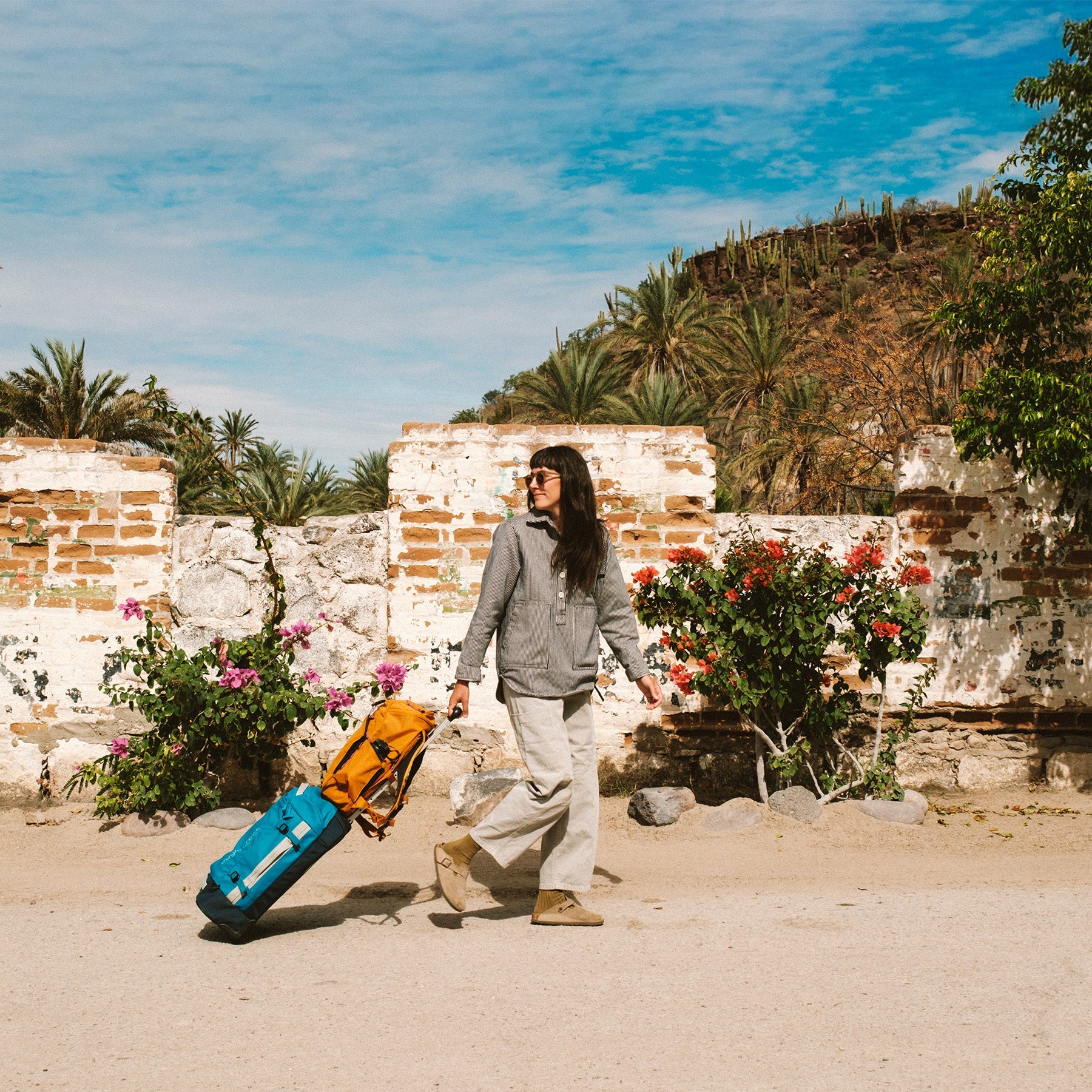 A woman rolls an orange travel backpack on top of a blue carry on duffel bag through the sand