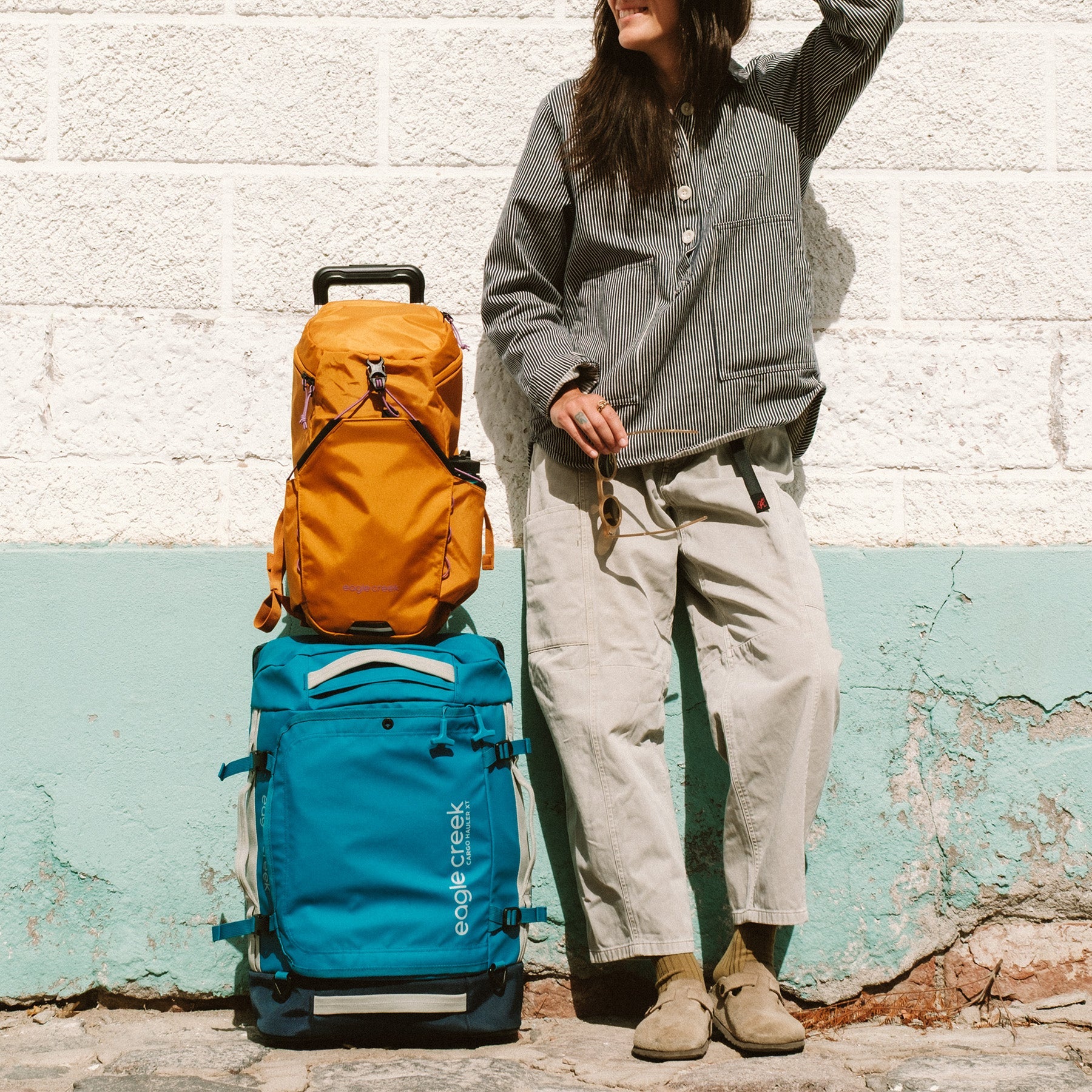 A woman stands with an orange travel backpack on top of a blue carry on duffel bag