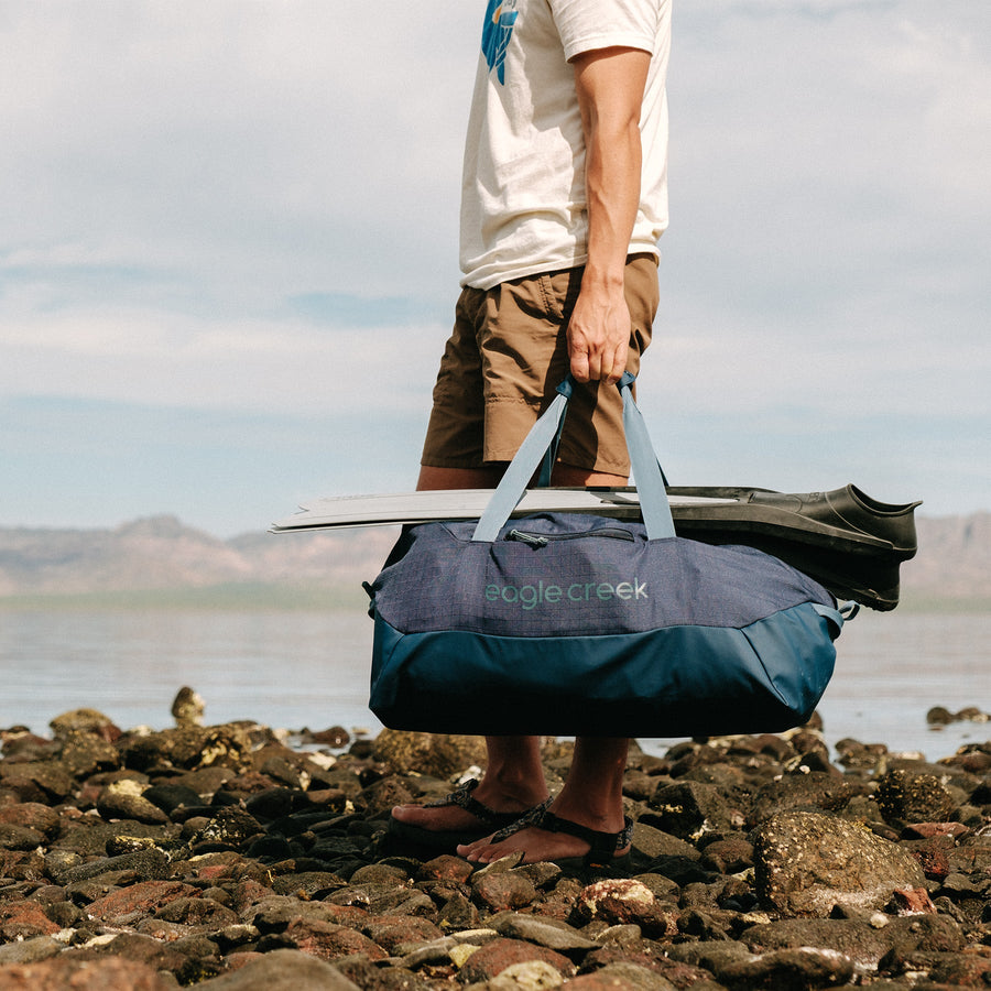 A person carrying a large blue duffle bag and a pair of flippers on a rocky beach
