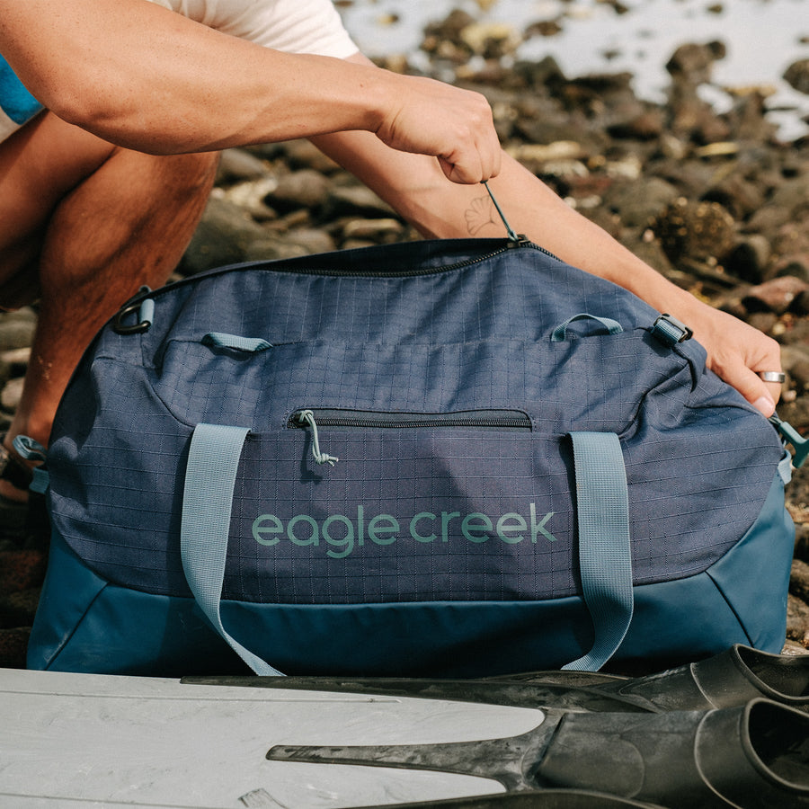 A person unzipping a navy blue duffle bag on a rocky beach