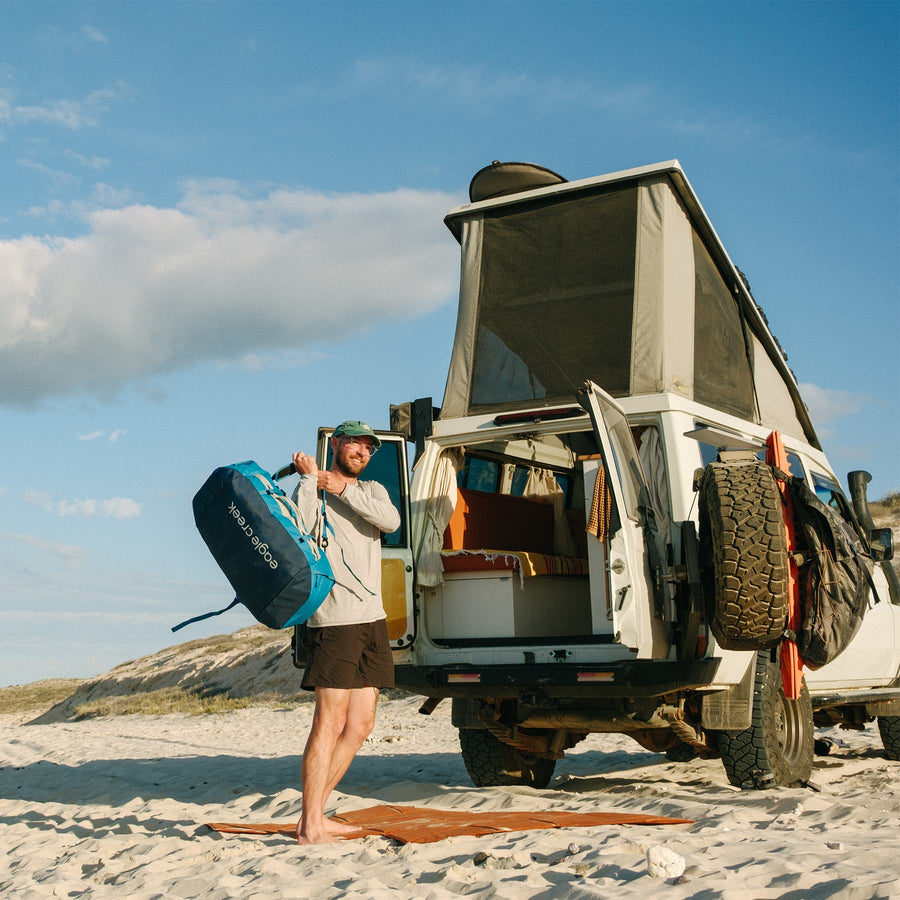 A man puts a blue duffle bag on his shoulder while standing next to a camper van on the beach